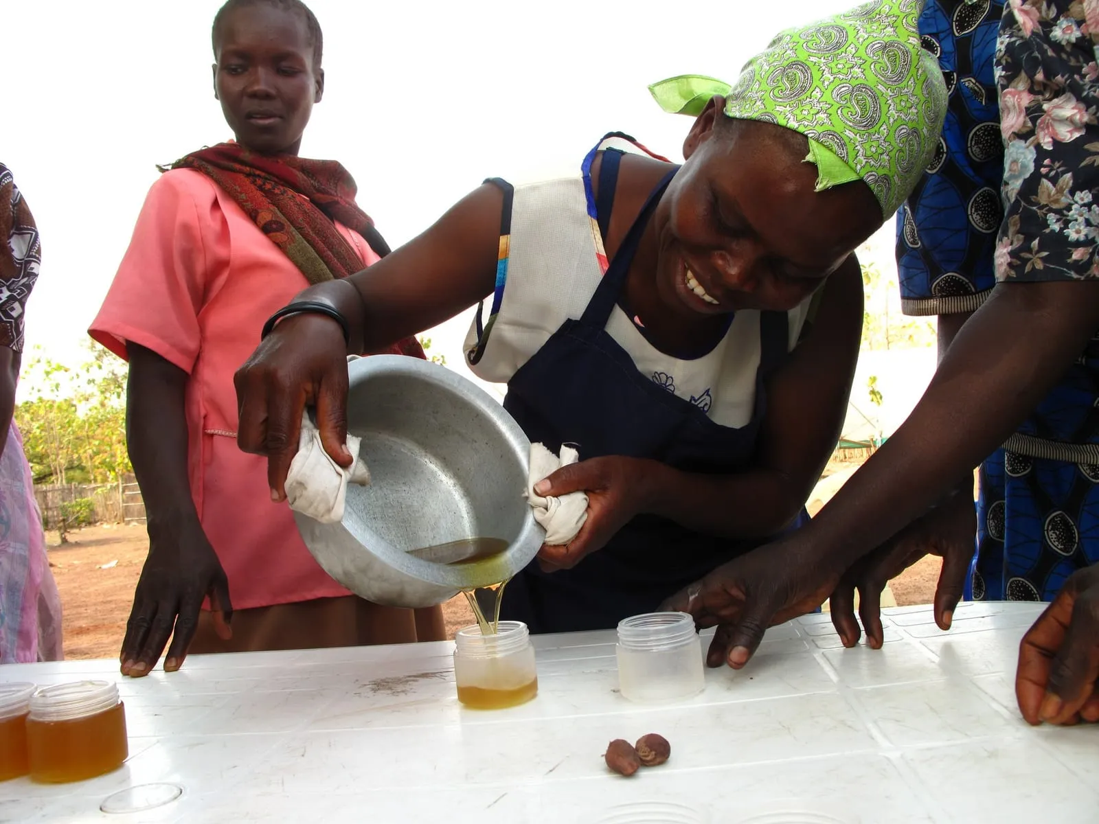 Women at a soap-making workshop pour a liquid from a metal bowl into small cups on a table.